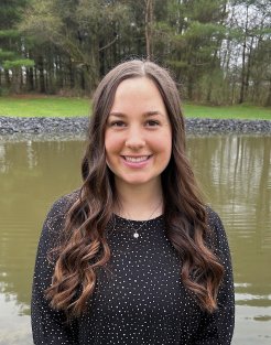 Sara Rachon, with long brown hair, stands in front of a body of water and smiles at the camera.