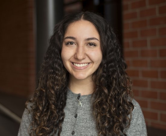 Olivia D’Ambro, with long, dark, curly hair and a gray shirt, stands in front of a brick wall and smiles.