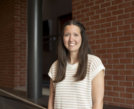 Kyla Reed, with long brown hair past her shoulders, wearing striped short sleeve shirt, stands in front of a brick wall and smiles.